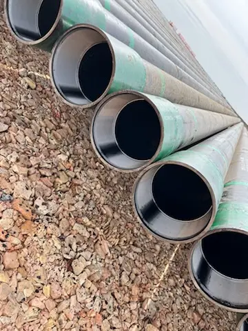 Stacked oilfield casing joints with green paint identification bands resting on gravel