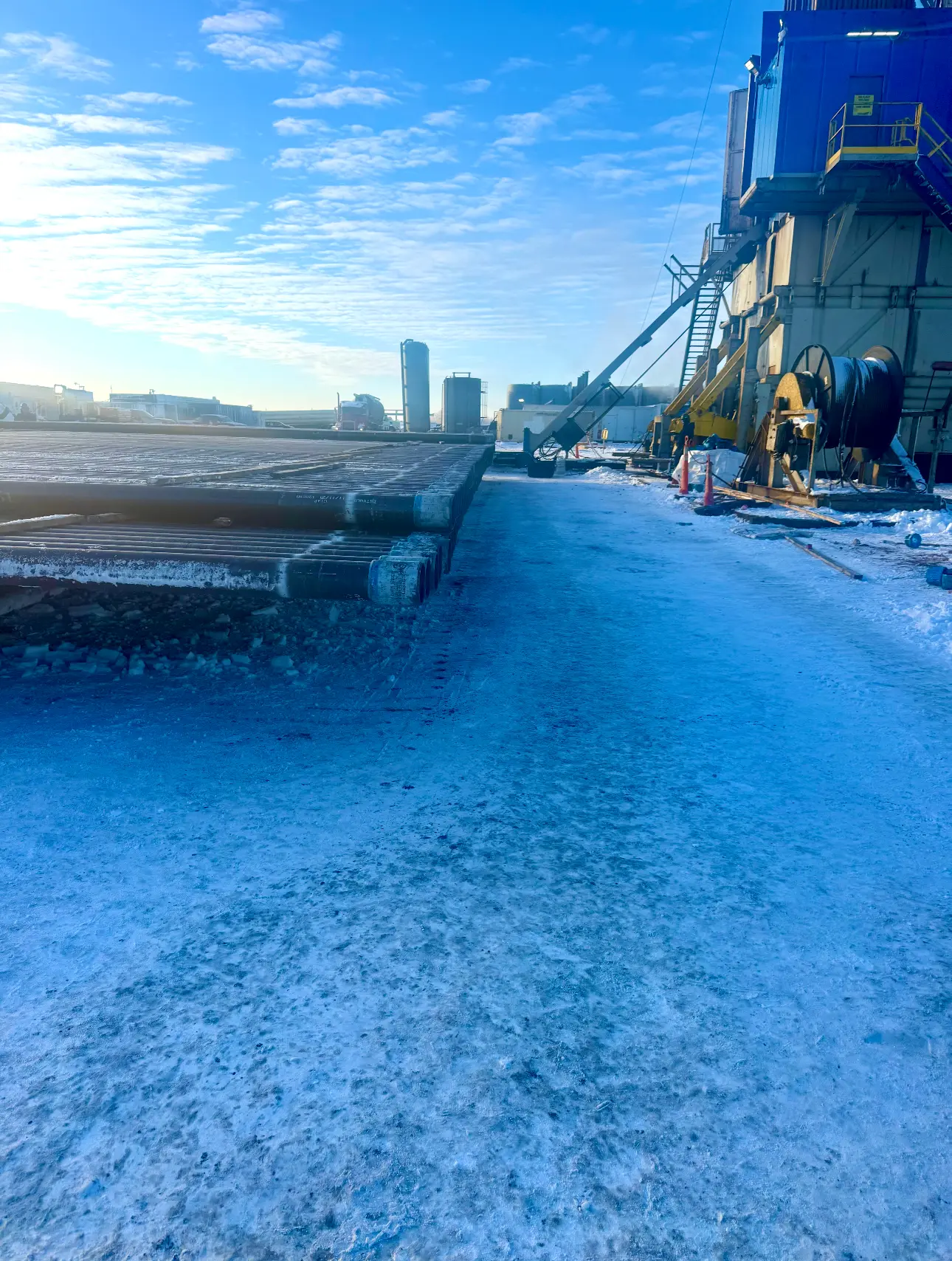 Stacked casing pipe on a frozen rig pad alongside a blue workover rig substructure in winter