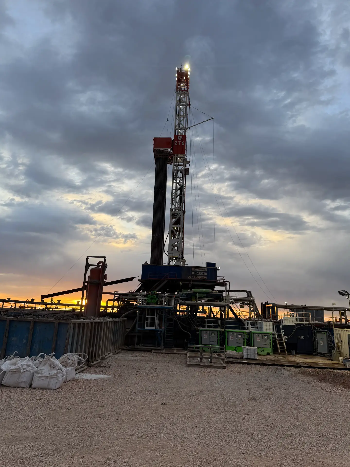 Drilling rig lit at dusk with stage solids control units and water tanks on a Bakken pad