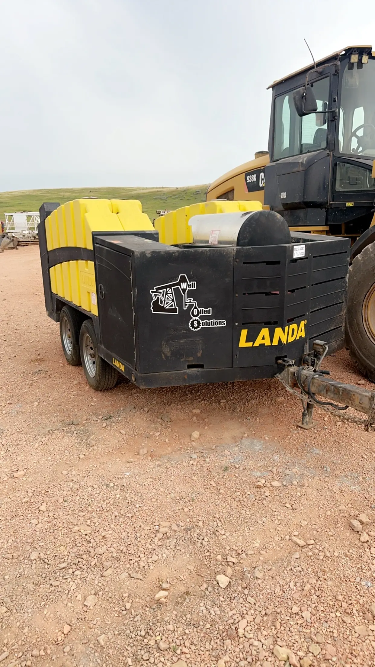 WOS Landa pressure-washer skid trailer parked next to a Cat 938K loader on a Bakken pad