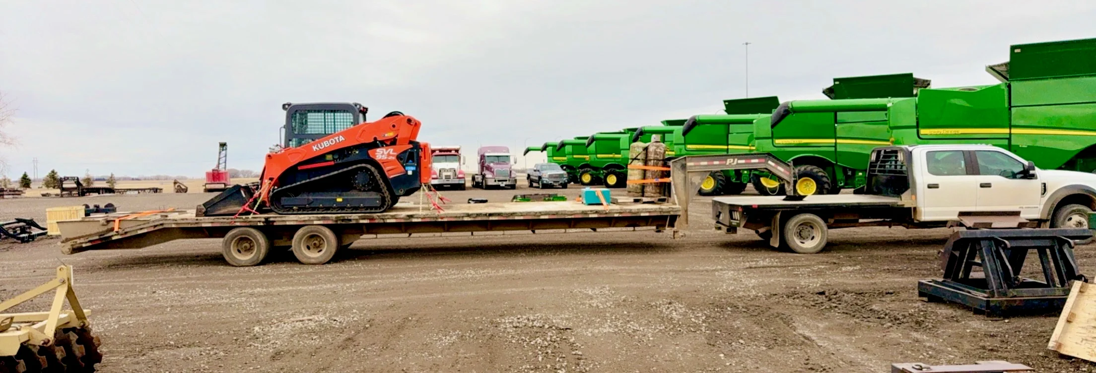Kubota skid steer loaded onto a WOS gooseneck flatbed at the yard alongside John Deere combines