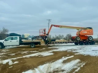 WOS flatbed truck hauling a JLG telescopic boom lift on a gooseneck trailer to the rig
