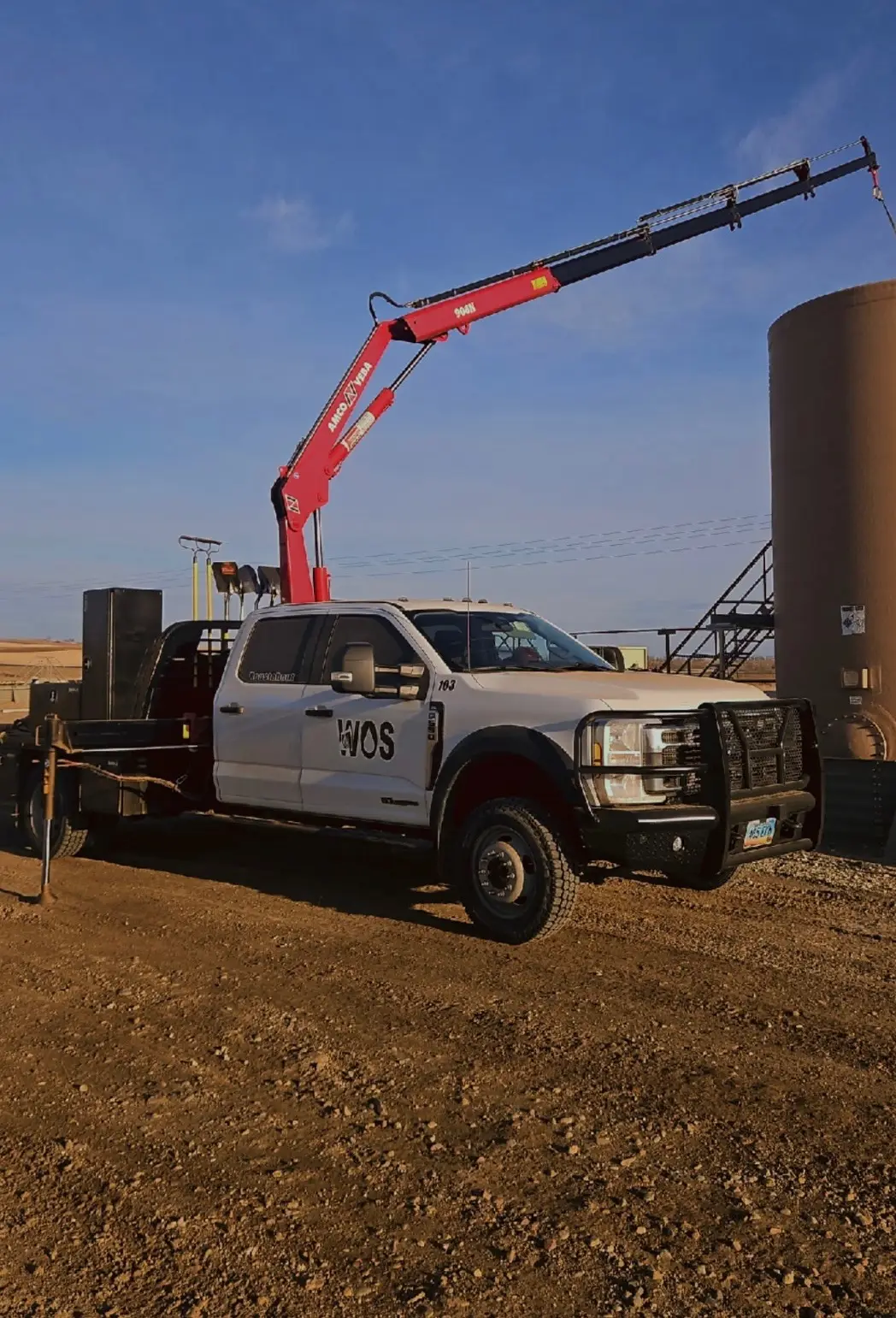 White WOS Ford F-550 with a red knuckle boom crane parked next to a production tank at sunset